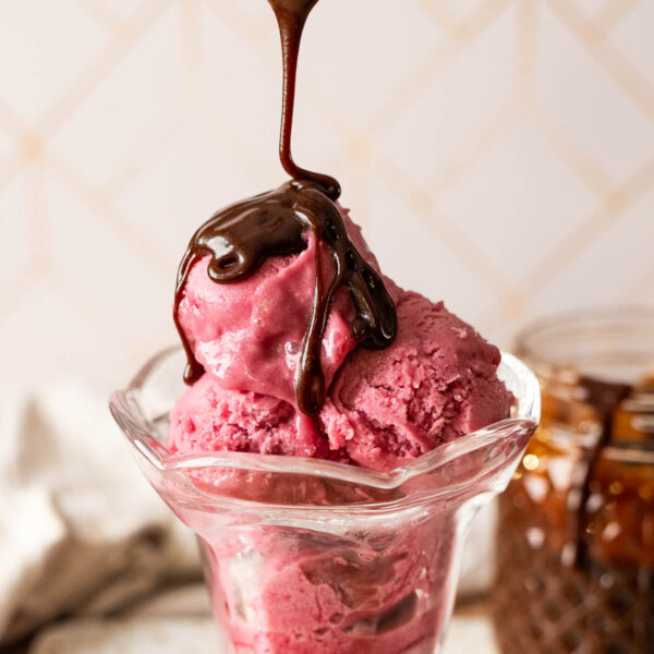 A glass dish holds scoops of berry ice cream as a rich hot fudge sauce is drizzled on top. A jar of sauce is visible in the background, making this gluten free treat irresistible.