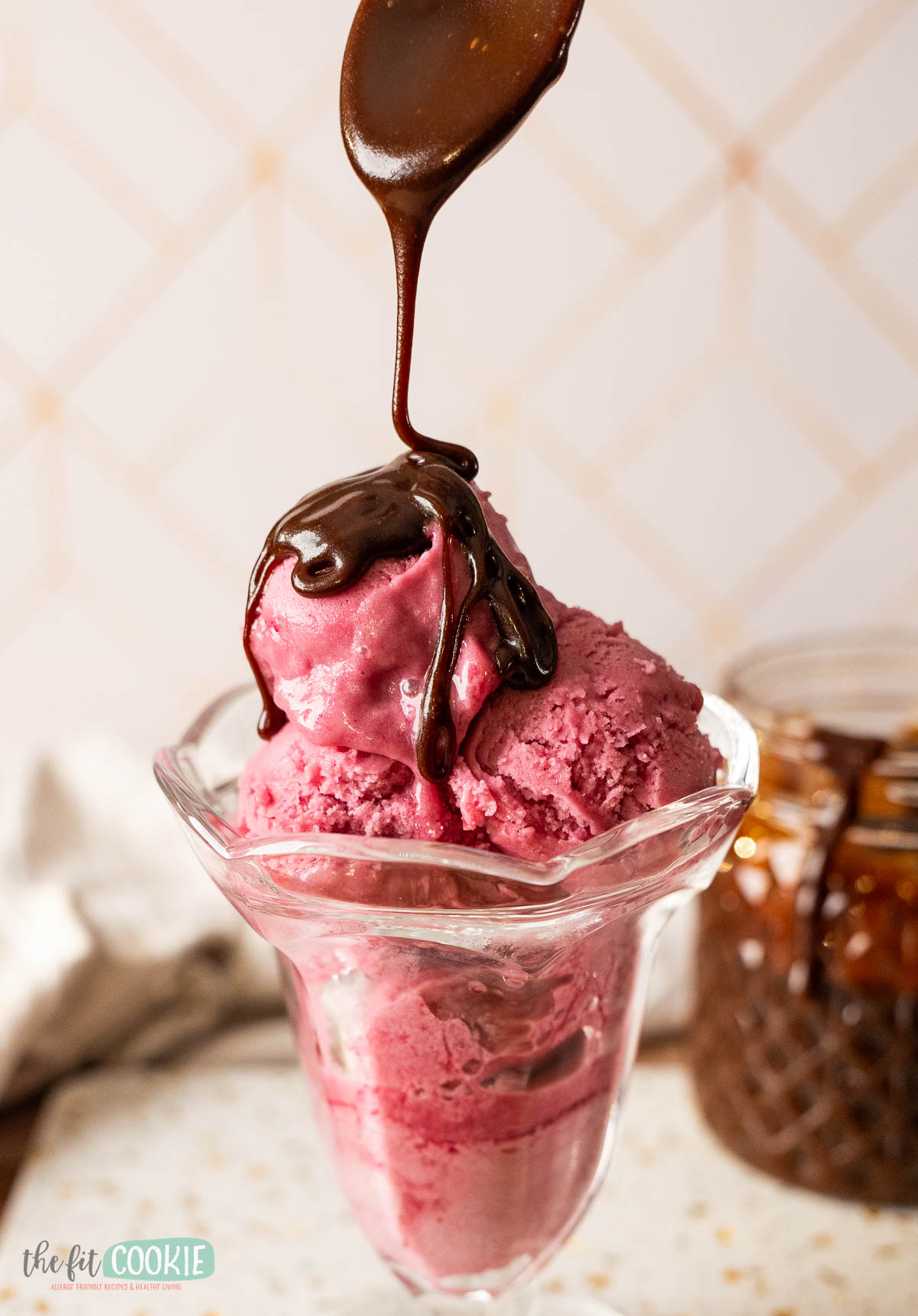 A glass dish holds scoops of berry ice cream as a rich hot fudge sauce is drizzled on top. A jar of sauce is visible in the background, making this gluten free treat irresistible.
