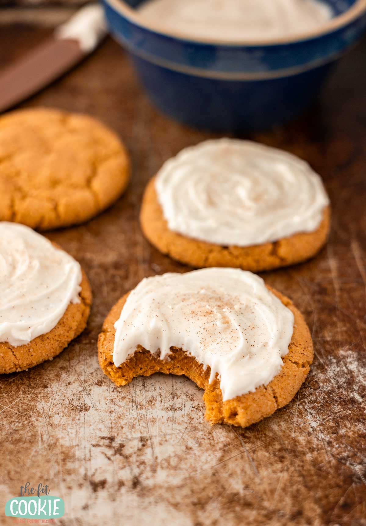 Three frosted gluten free pumpkin snickerdoodles sit on a sheet pan, one with a bite taken out. A bowl of frosting and a spatula are visible in the background.