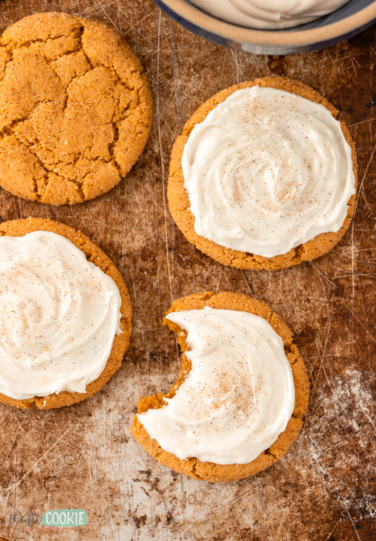 Four pumpkin snickerdoodles rest on a worn baking sheet, three with dairy free cream cheese frosting and a sprinkle of cinnamon, one with a bite taken out; a bowl of frosting visible in the corner.